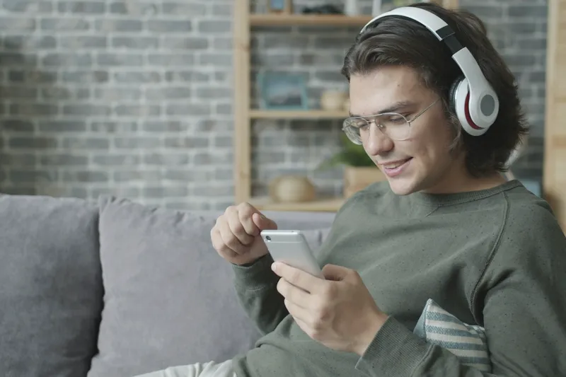 A young man with headphones on a sofa, smiling at his phone.