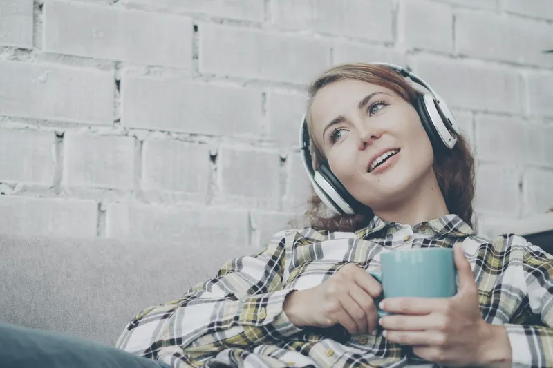 A woman resting on a sofa with headphones and a mug.